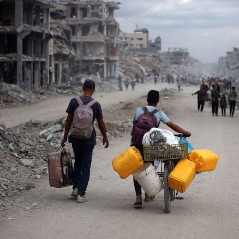 People walk through a street filled with rubble and destroyed buildings; one person pushes a bicycle loaded with yellow water containers and supplies. The scene depicts devastation and displacement.