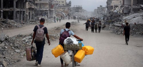 People walk through a street filled with rubble and destroyed buildings; one person pushes a bicycle loaded with yellow water containers and supplies. The scene depicts devastation and displacement.