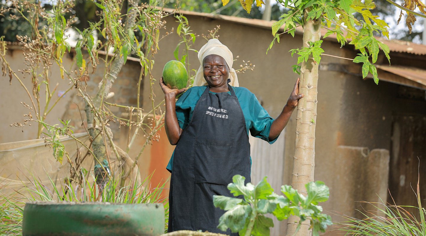 a woman stands in a garden, holding up a large recently-harvested fruit and smiling.