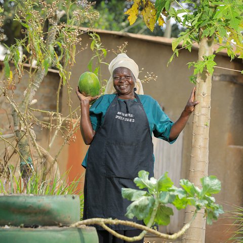 a woman stands in a garden, holding up a large recently-harvested fruit and smiling.