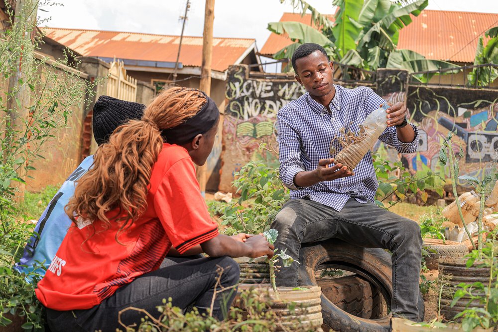 a man, sat in a garden, showing a plastic bottle with spices growing inside to two others.