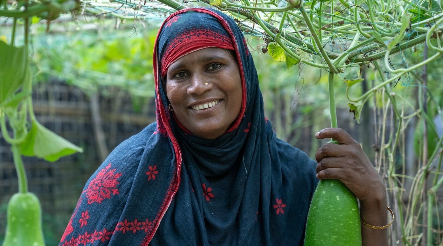 A woman in Cox's Bazar smiling and posing with the produce she is growing in a kitchen garden.