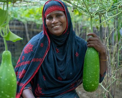 A woman in Cox's Bazar smiling and posing with the produce she is growing in a kitchen garden.