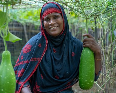 A woman in Cox's Bazar smiling and posing with the produce she is growing in a kitchen garden.