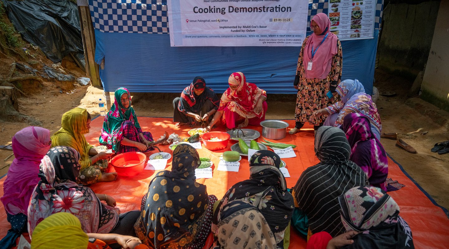A group of women are sat on a large orange rug with bowls and sheets of paper participating in a practical cooking demonstration.
