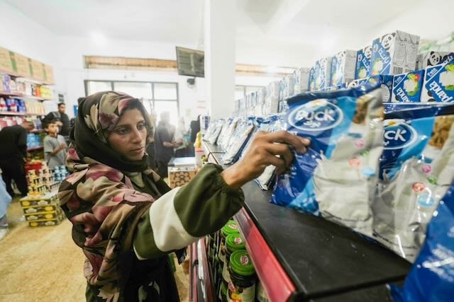 Close-up of a woman reaching for packets of dairy product from a well-stocked shelf as other shoppers queue in the background.