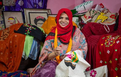 A smiling woman in a red head scarf stitching a jute embroidery, with an selection of her hand-stitched pillows and blankets in the background.