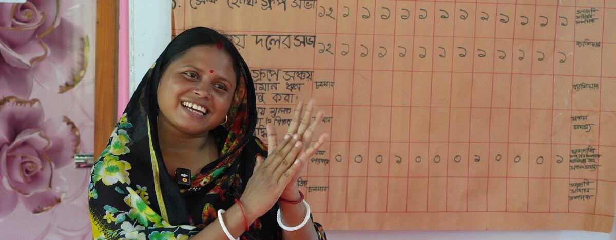 A person stands in front of a handwritten wall chart, clapping, in a classroom or community meeting space. The chart behind them shows rows and columns of text and numbers in Bangla.