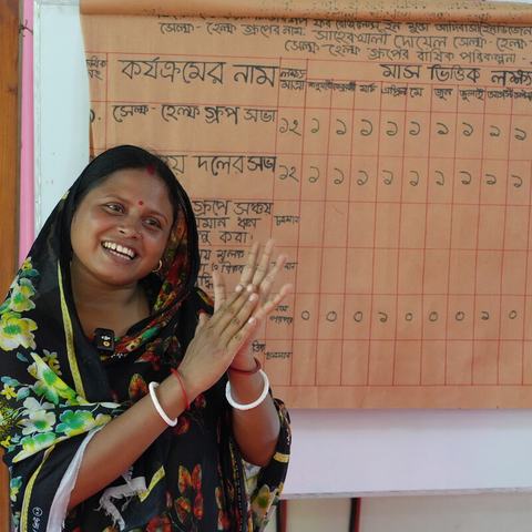 A person stands in front of a handwritten wall chart, clapping, in a classroom or community meeting space. The chart behind them shows rows and columns of text and numbers in Bangla.