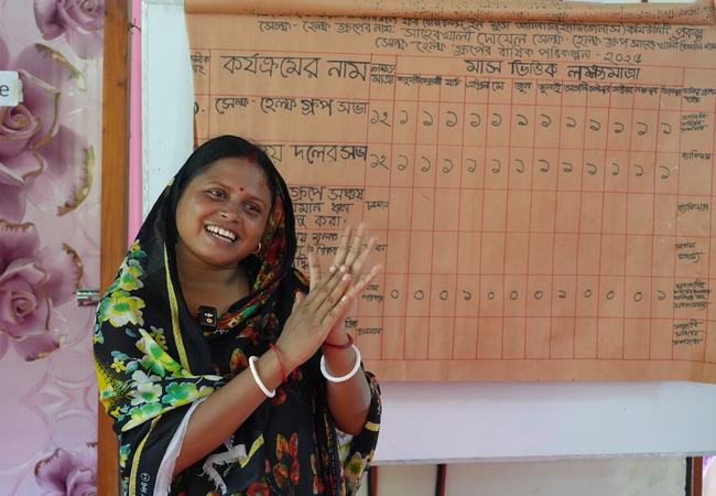 A person stands in front of a handwritten wall chart, clapping, in a classroom or community meeting space. The chart behind them shows rows and columns of text and numbers in Bangla.