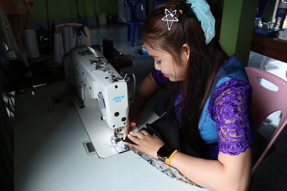 a woman sat at a sewing table, where she is using a sewing machine to stitch a patterned garment.