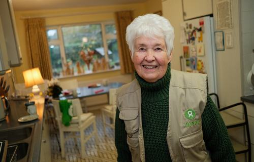 A smiling woman in an Oxfam-branded jacket and a green jumper stands in her kitchen.