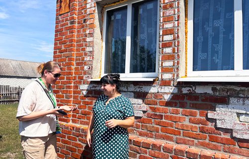 Two women stand outside a red brick building, talking near a set of repaired windows in bright sunlight. One holds a notepad or device while speaking with the other.
