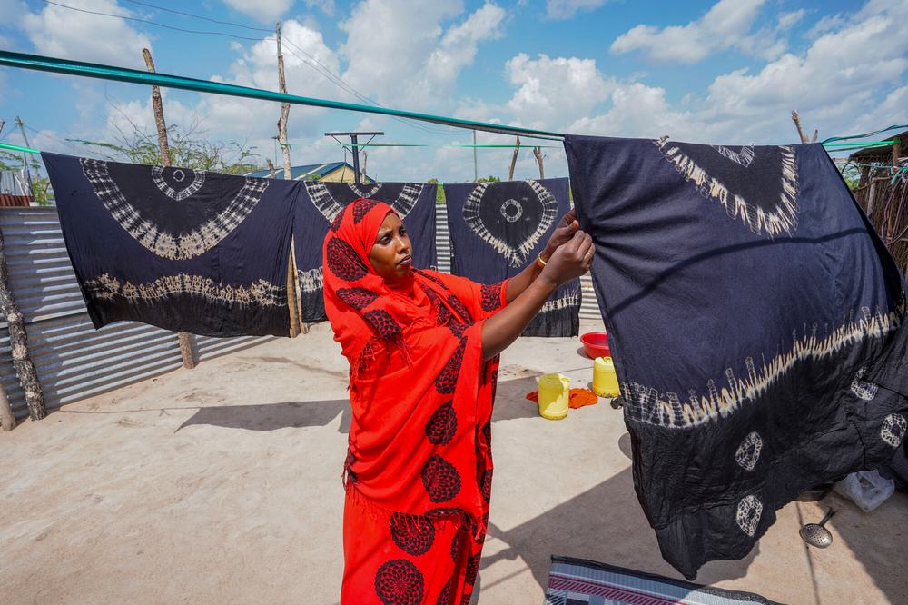 a woman stands by a clothesline on which several colourful tie-dyed garments are hanging.