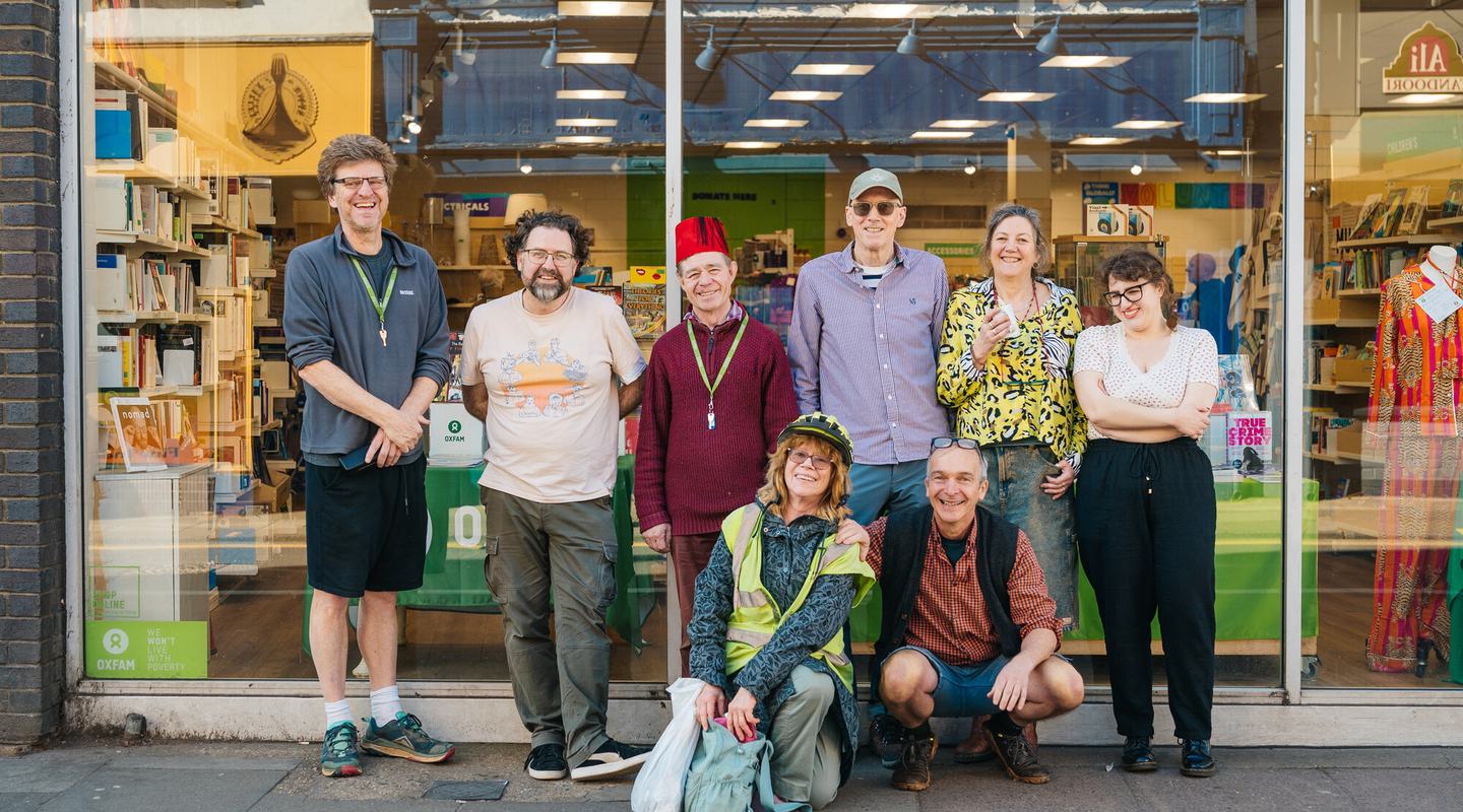 Group of eight people posing together on a pavement outside an Oxfam charity shop with large glass windows and books visible inside.