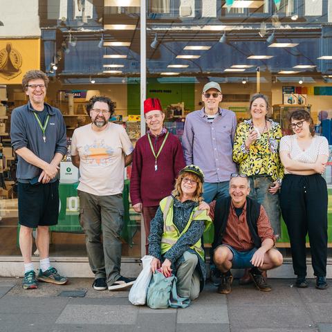 Group of eight people posing together on a pavement outside an Oxfam charity shop with large glass windows and books visible inside.