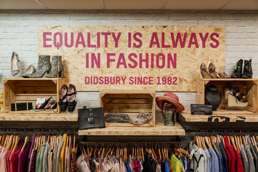 Oxfam Didsbury shop, surrounded by clothing rails a poster reads "Equality is always in fashion. Didsbury Since 1982"