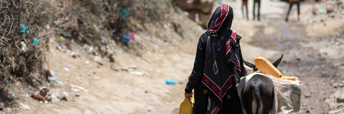 Person walking beside a donkey carrying full water containers along a dry, dusty track.