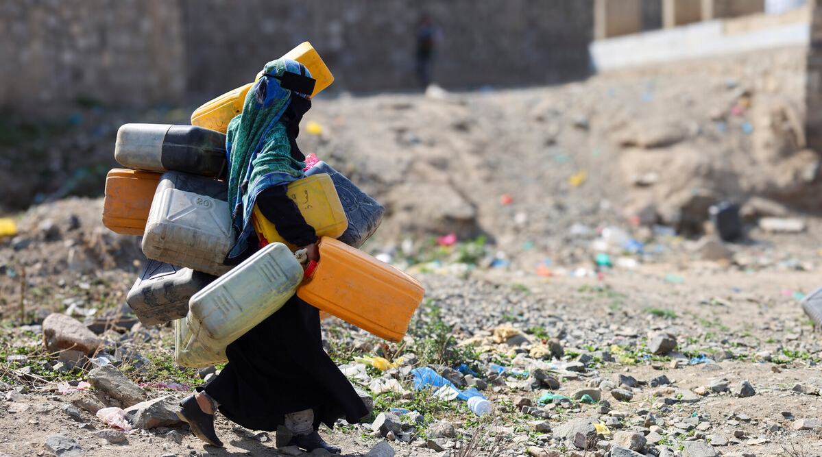 A woman in the city of Taiz, Yemen carrying lots of large water containers in her hands, under her arms and on her back walking across rubble.