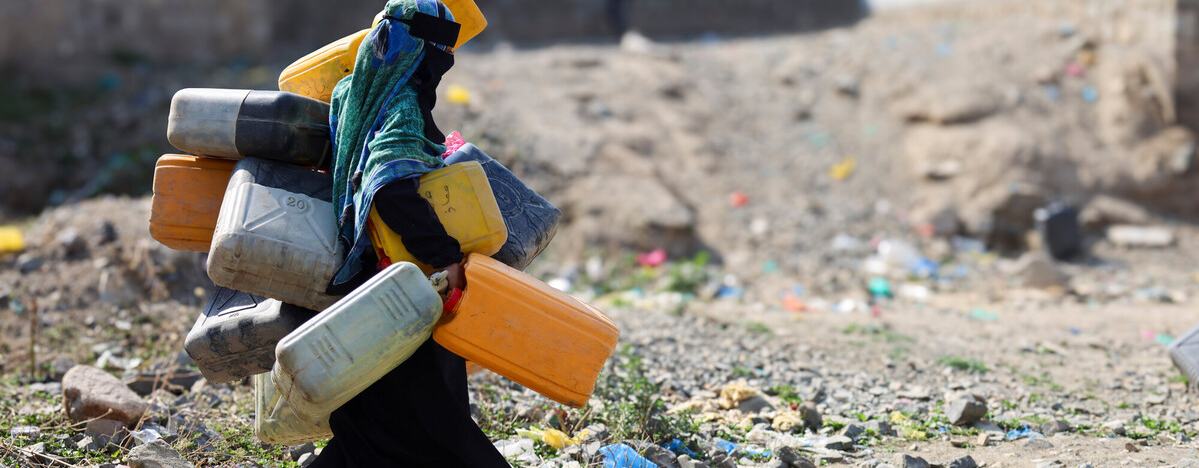 A woman in the city of Taiz, Yemen carrying lots of large water containers in her hands, under her arms and on her back walking across rubble.