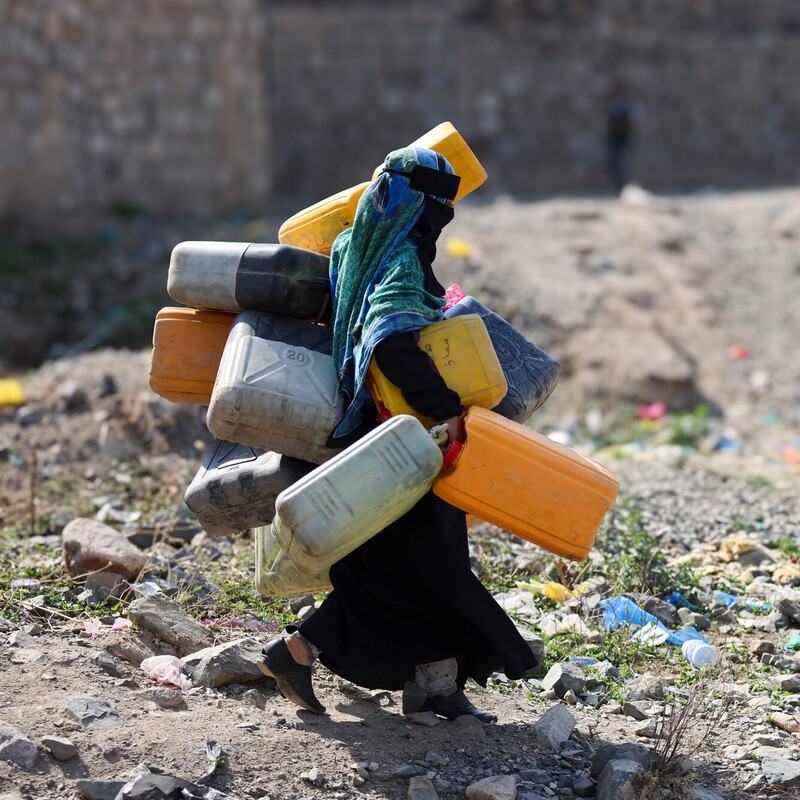 A woman in the city of Taiz, Yemen carrying lots of large water containers in her hands, under her arms and on her back walking across rubble.