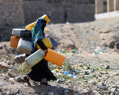 A woman in the city of Taiz, Yemen carrying lots of large water containers in her hands, under her arms and on her back walking across rubble.