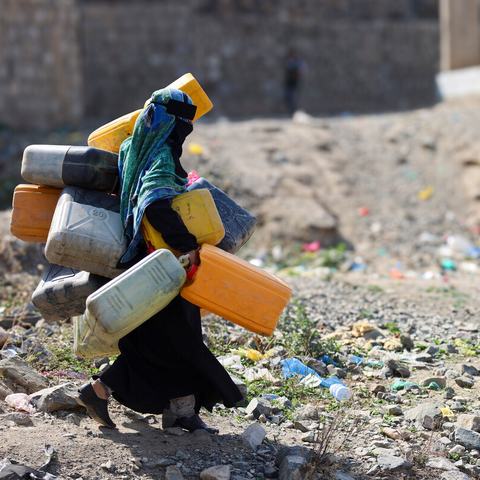 A woman in the city of Taiz, Yemen carrying lots of large water containers in her hands, under her arms and on her back walking across rubble.