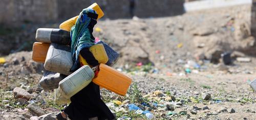 A woman in the city of Taiz, Yemen carrying lots of large water containers in her hands, under her arms and on her back walking across rubble.