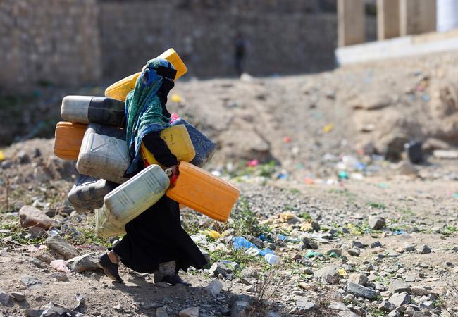 A woman in the city of Taiz, Yemen carrying lots of large water containers in her hands, under her arms and on her back walking across rubble.