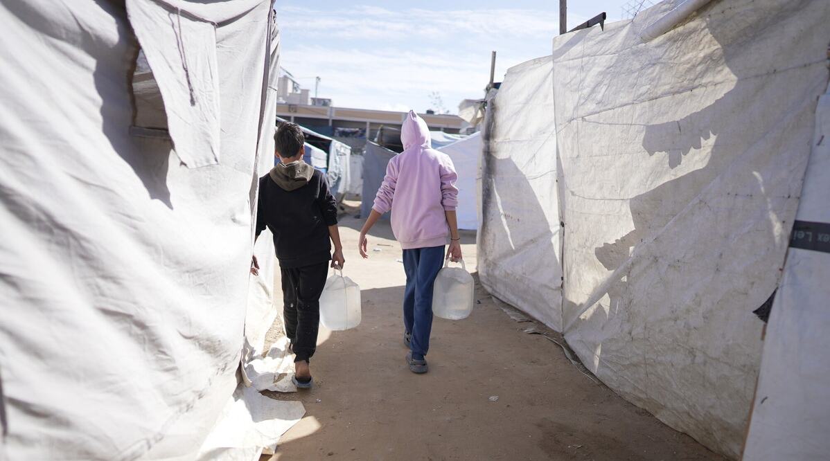Two people carrying water containers walk between rows of temporary shelters in a crowded camp.