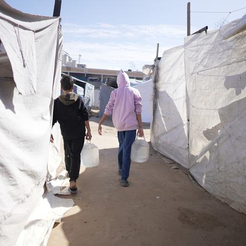 Two people carrying water containers walk between rows of temporary shelters in a crowded camp.