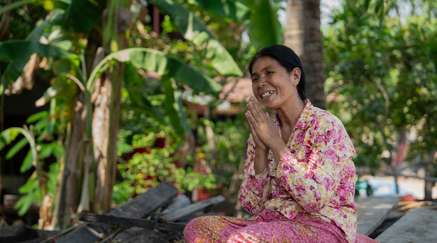 A smiling woman holds her hands together as she sits on a pile of wood in a camp for displaced people in Cambodia.