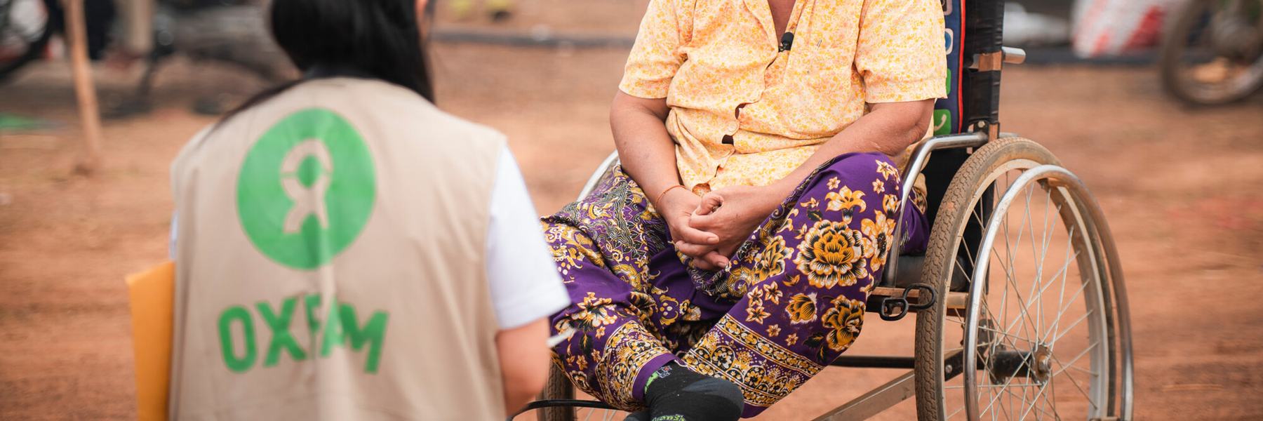 A woman in an Oxfam-branded vest speaks to a woman in a wheelchair in Chong Kal camp, Cambodia.