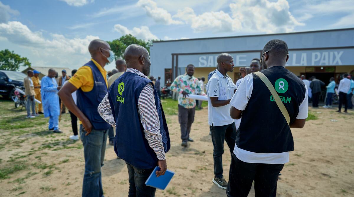 A group of people wearing Oxfam vests stand and talk in a busy outdoor setting near a building marked “Salle de la Paix.”