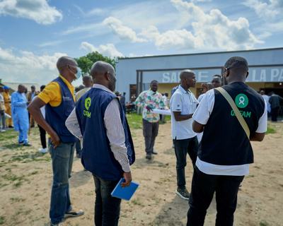 A group of people wearing Oxfam vests stand and talk in a busy outdoor setting near a building marked “Salle de la Paix.”