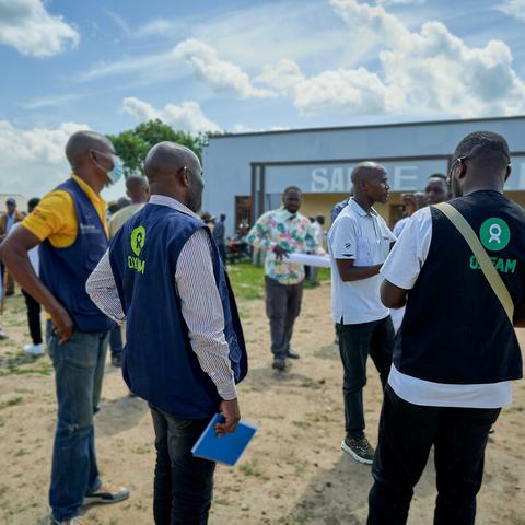 A group of people wearing Oxfam vests stand and talk in a busy outdoor setting near a building marked “Salle de la Paix.”