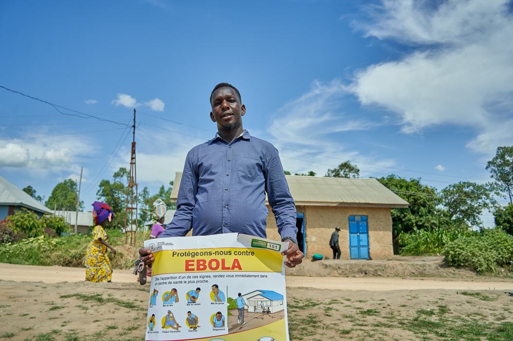 Man standing outside under blue sky, holding an awareness sign about Ebola