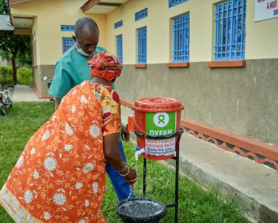 A male health worker looks on as a woman uses a water bucket tap in the grounds of the Rwampara General Referral Hospital, Mongbwalu, DR Congo.