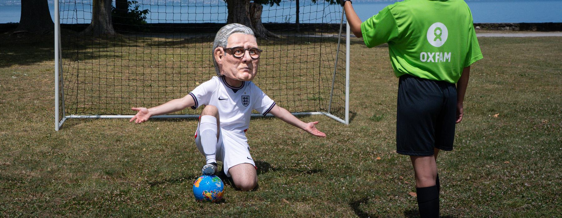 Oxfam campaigner with a Keir Starmer' Big Head in a football strip kneeling inside a goal, being given a red card. A banner reads world aid cuts.
