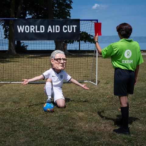 Oxfam campaigner with a Keir Starmer' Big Head in a football strip kneeling inside a goal, being given a red card. A banner reads world aid cuts.