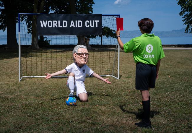 Oxfam campaigner with a Keir Starmer' Big Head in a football strip kneeling inside a goal, being given a red card. A banner reads world aid cuts.