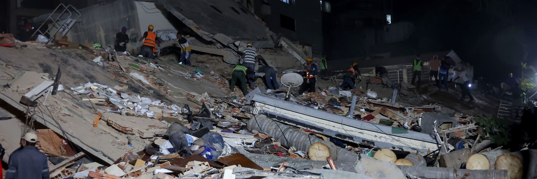 Rescue workers search through the rubble of a collapsed building at night, illuminated by floodlights. Large piles of concrete, debris, and damaged household items cover the site, with apartment buildings visible in the background.