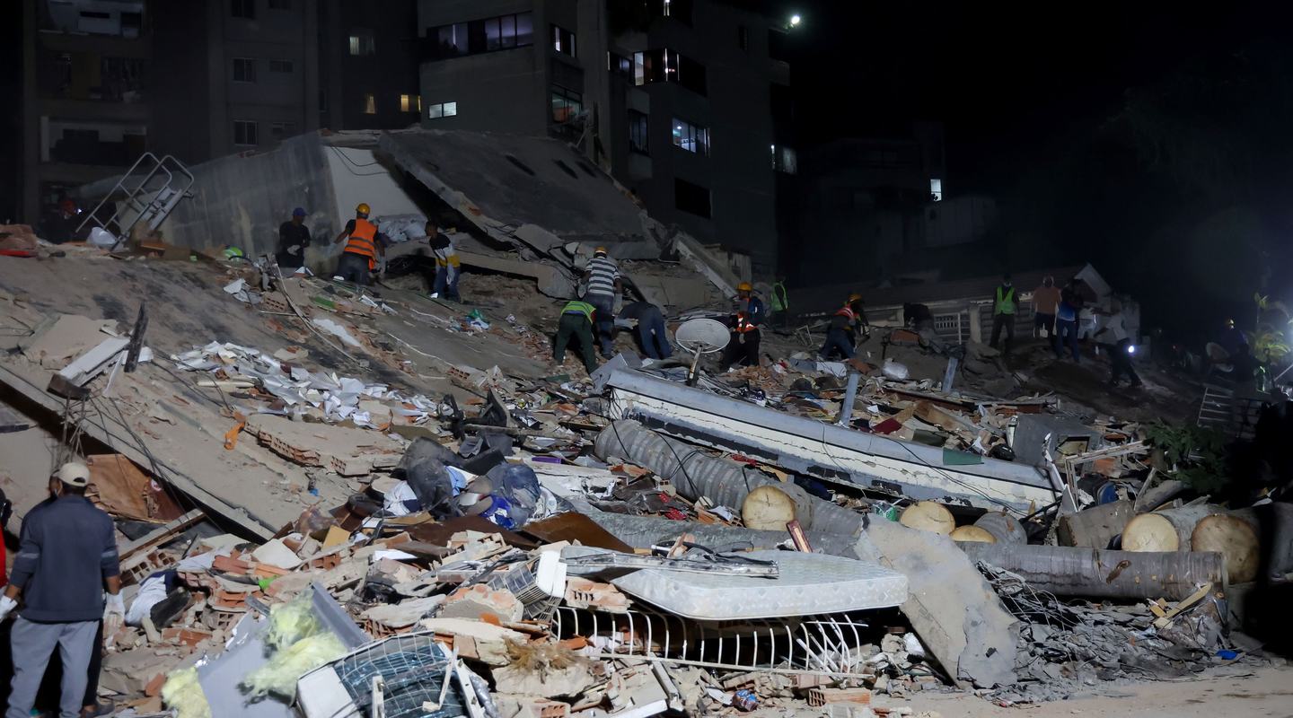 Rescue workers search through the rubble of a collapsed building at night, illuminated by floodlights. Large piles of concrete, debris, and damaged household items cover the site, with apartment buildings visible in the background.