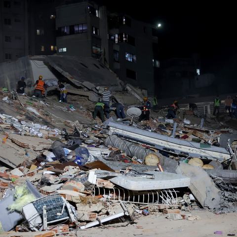 Rescue workers search through the rubble of a collapsed building at night, illuminated by floodlights. Large piles of concrete, debris, and damaged household items cover the site, with apartment buildings visible in the background.