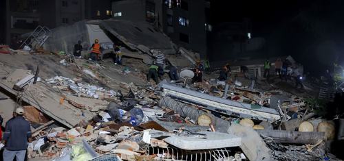 Rescue workers search through the rubble of a collapsed building at night, illuminated by floodlights. Large piles of concrete, debris, and damaged household items cover the site, with apartment buildings visible in the background.