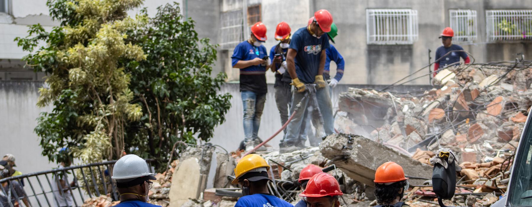Rescue teams are searching for victims in the San Bernardino area of Caracas. Men in hard hats stand on top of rubble with equipment.