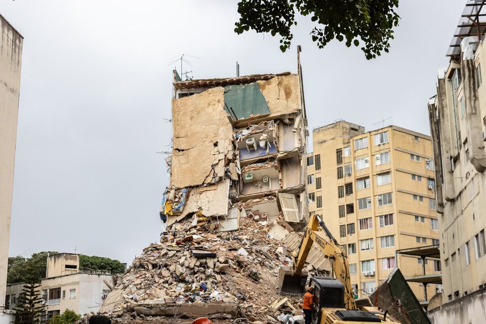 A severely damaged multi-storey apartment building stands partially collapsed amid a large pile of rubble, with an excavator clearing debris in an urban area.
