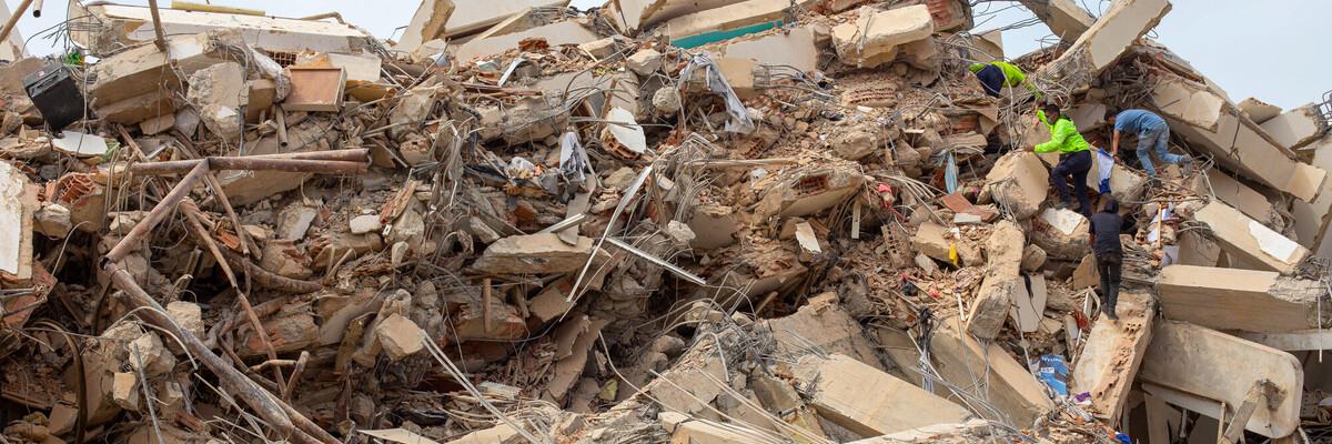 Rescue workers climb over the remains of a collapsed concrete structure, searching through piles of rubble, broken beams and exposed steel reinforcement.