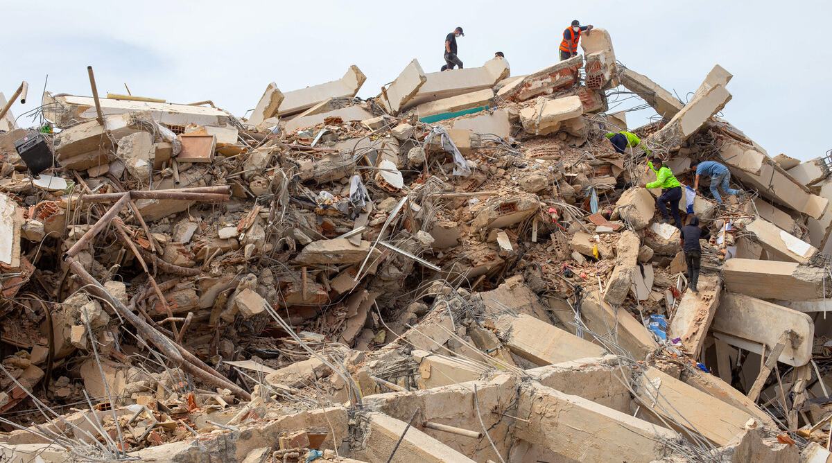 Rescue workers climb over the remains of a collapsed concrete structure, searching through piles of rubble, broken beams and exposed steel reinforcement.