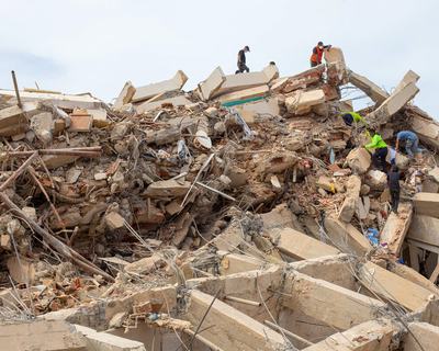 Rescue workers climb over the remains of a collapsed concrete structure, searching through piles of rubble, broken beams and exposed steel reinforcement.
